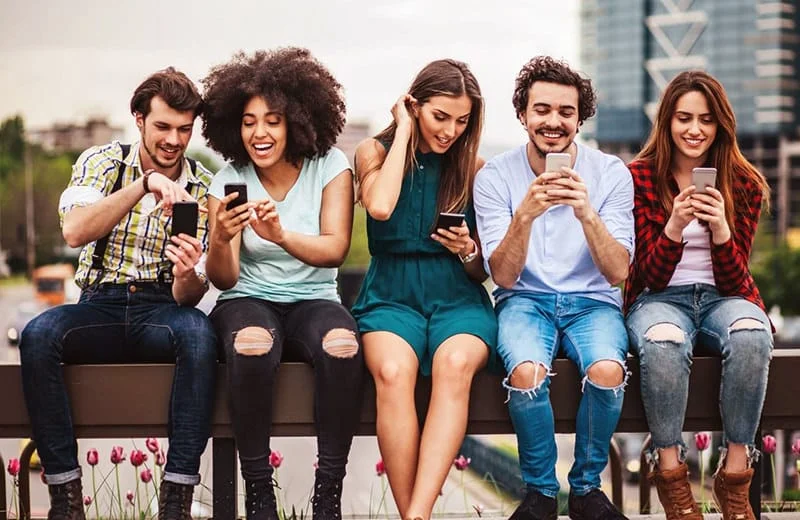 22-trucos-redes-sociales-eduweb Menores de edad usando sus teléfonos móviles en un parque urbano con edificios de fondo, mostrando interés en tecnología y comunicación digital.